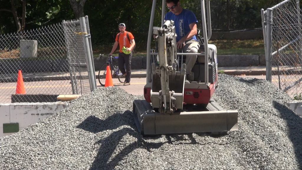 Placetailor director of development, Declan Keefe, arrives at the job site by bicycle, as company operations manager, Evan Smith, moves a fresh load of gravel onto the foundation slab pad using a skid-steer mini-excavator. Educated as architects but experienced in construction, Keefe and Smith are co-owners of the company, and the only members remaining from Placetailor's original group of founders.