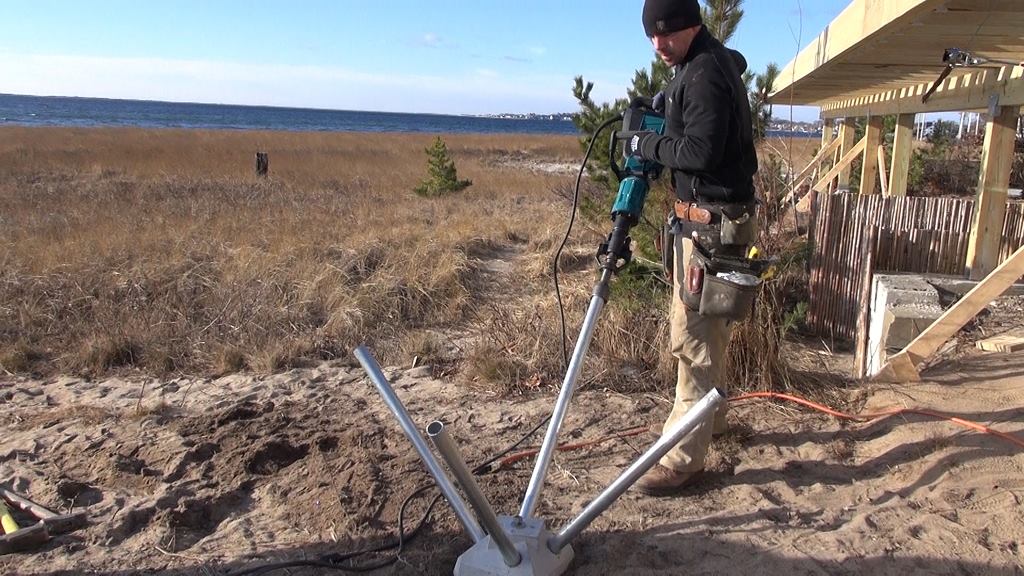 Mike Horgan uses an electric jackhammer to drive pins through a Diamond Pier block (above). With access limited through the sensitive beach and dunes (below), Horgan was not allowed to bring digging equipment on site. The Diamond Pier allowed Horgan to set code-compliant footings using just a shovel and an electric jackhammer. Working his way around each Diamond Pier and driving the pins with the jackhammer (bottom) took about ten minutes per pier.