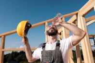 The man builder sits on the edge of the roof of the frame house, in a yellow helmet and gray overalls and a white T-shirt. Sweats and pours water on the head from the bottle. Hard work and rest.