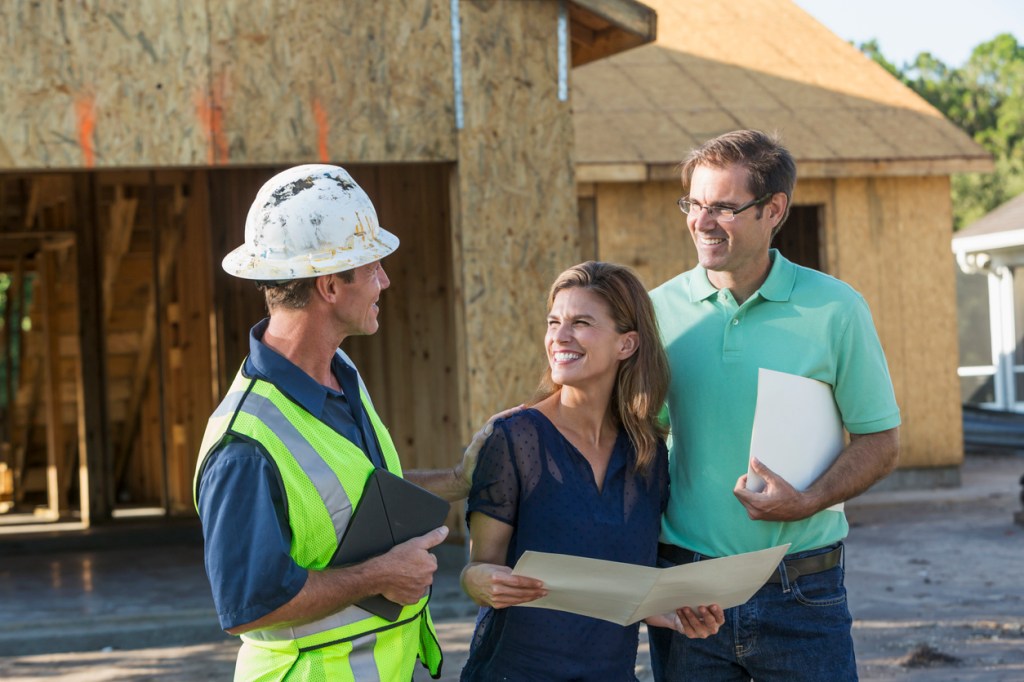 A mature couple standing with a contractor or construction worker outside their new home under construction.  They are talking and holding folders.  The worker who is wearing a hardhat and safety vest is holding a digital tablet.