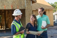 A mature couple standing with a contractor or construction worker outside their new home under construction.  They are talking and holding folders.  The worker who is wearing a hardhat and safety vest is holding a digital tablet.