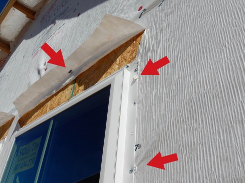 This photo shows several problems, identified with arrows: The top arrow shows where a nail is used to hold the head flap up, creating two unnecessary holes in the WRB. The head flap should be temporarily held in place with tape. The middle arrow shows sealant squeeze-out left to harden like a worm. Any SAF applied over this will not perform well. The lowest arrow shows where a cap fastener is placed too close to the nail fin. As a rule of thumb, fasteners should be no closer than “three fingers” from the rough opening; Tyvek says 6 inches.