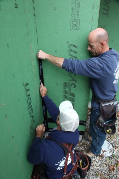 One crewmember keeps the length of flashing tape folded while the other adheres the first side, pushing the tape tight into the corner with a plastic spreader. After the first side is adhered, the adjacent side can be pressed into place.