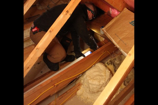 A crew member replaces a length of 4-inch flex duct run from a bath fan with 4-inch rigid PVC piping. The PVC duct sloped 1/4 inch per foot toward the soffit vent. In the foreground, a recessed light is air-sealed with a site-built OSB cover foamed in place.