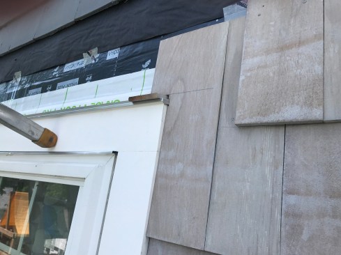 The author steps the shingles around the corner of the window trim, using a scrap shingle butt to maintain a drainage gap above the drip cap. 