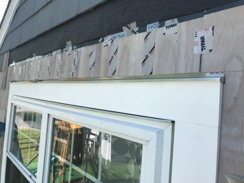 The author measures and cuts all the shingles on the ground for above the window. He tapes the shingles together temporarily so that he can bring them up the ladder and install them as a single unit. 