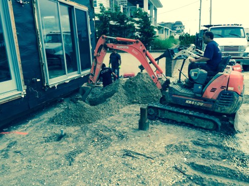 To make room for  driving the pier sections under the overhanging wall, a bucket excavates a hole at each pier position. Around 4 feet of vertical clearance is needed to drive the piles.