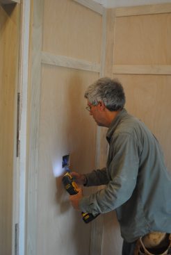 As with the receptacles on the long wall, the author lays out the box opening on the panel, cuts a hole in the middle to verify the box location, and then carefully cuts the box out with a multi-tool. The electrician will add box extenders to cover the combustible edges of the panel material. 
