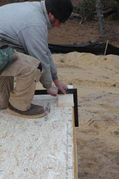 The first layout task is to establish a base line or reference line for locating the rest of the layout lines. Here the lead carpenter uses a layout block made from plate stock and a framing square to align the layout with the deck framing.