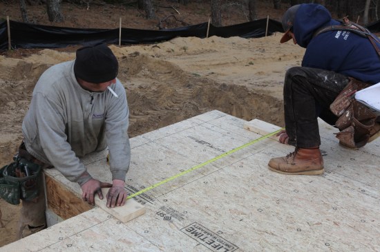 To lay out the fireplace walls, both crew members use their layout blocks to hold and mark the measurement.