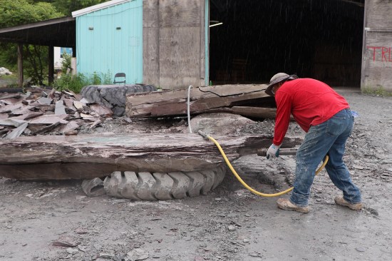 A worker uses an air hammer to split a slate block down to 6-inch-or-less thicknesses in order to fit under the cutting saw arbor. Upon excavation, slate has to be managed carefully. It has a certain amount of moisture content and its cleaving planes can be ruined if the stone freezes or it lays out in the sun too long and dries out; wind is also a factor in freezing and drying. Each fall, slate blocks are stacked in the storage building beyond to maintain production in winter.