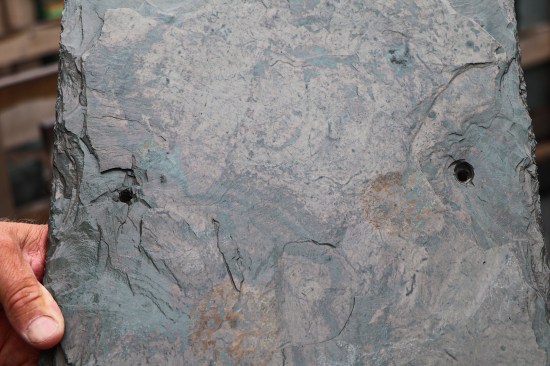 The front of a 1-inch-thick unfading mottled green and purple slate after hammer-drilling the punch holes. Pricing for a square roof slate ranges widely due to color selection and thickness. The least expensive slate is roughly $400 a square, while the most expensive goes for over $2,000 a square.