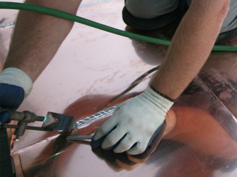 Soldering is two-handed work: One hand works the iron; the other controls the solder stick. While holding the iron at a shallow angle with the tip against the metal, press the solder stick against one facet of the tip until a drop of melted solder forms, then use the iron to spread the drop across the seam. Each drop adds another layer, and the heat of the iron on the copper draws the solder into the seam to make a watertight seal. For soldering tips, see the “Soldering Gutters” video and “Soldering Seams” slideshow.