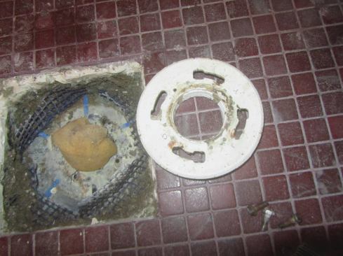Second, someone had shimmed up the clamp ring with blue plastic shims spaced around the perimeter. This prevented the ring from firmly clamping the liner down onto the drain body below. Our best guess is that the tile guys who installed this drain turned the ring over in an effort to extend the thickness to account for a second layer of old tile underneath. But they knew that the upside-down ring needed weeps, so they tried to make a weep passage by shimming the clamp ring up a bit. Big mistake.