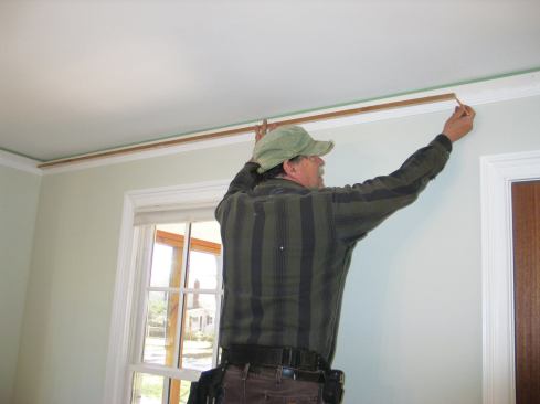 I cut a strip of wood exactly 100 inches long, butt it into one corner, and mark the end on the wall. Then I measure from the mark to the other corner with a metal ruler, adding 100 to that measurement to find the total length.