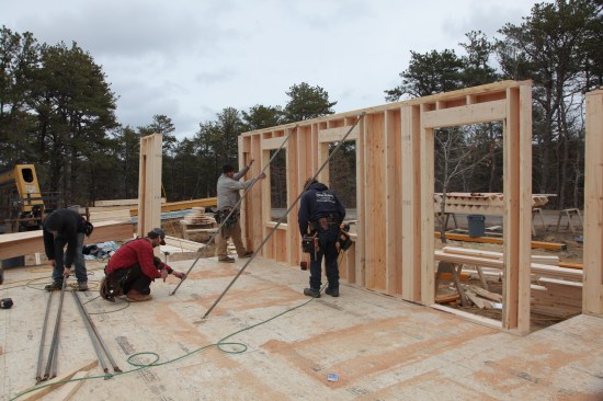 When the walls are standing and the bottom plate is nailed to the deck, a crew member holds the wall steady while adjustable braces are attached. A laser level on the bottom plate is used to gauge the wall plumb.