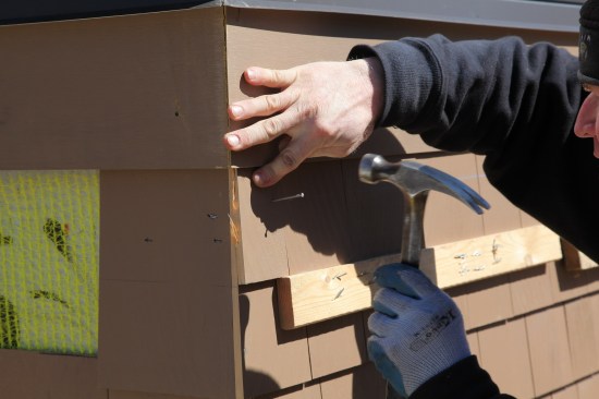 Using the 1-by ledger as guide, the corner shingle for the top course is nailed on. It will be smoothed with a plane before the adjacent shingle is added.