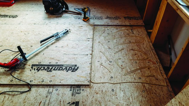 The crew installed the second layer of subflooring perpendicular to the first with joints staggered in both directions. They made sure to maintain 1/8-inch clearance between the sheets on all sides.