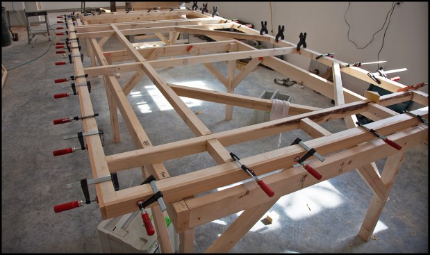 The inner grid incorporated a continuous vertical grid member, which provided solid blocking for the exterior-facing T1-11 skin (a 1-foot-wide add-on panel of T1-11 was needed to increase the T1-11’s width to 5 feet — see next slide). Also, a rough opening for the transom window, diagonal bracing, and blocking at the heavy-duty strap hinge locations (four per door) were incorporated. Here (photo, above), the cedar weather edge strips are glued up and clamped in place. 