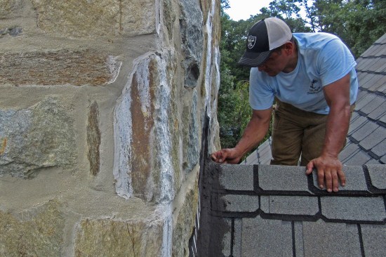 An M & J trade partner inspects the chimney’s base; roofing cement covers its shingle-to-base-flashing juncture.