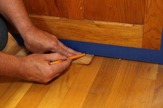 Next, he scribes to fit the contour of the floor and the wall, using a pencil and a scrap of wood that matches in thickness the widest gap he shimmed (he didn’t have dividers on hand for this job).