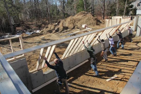 Working together, the entire crew picks up the long wall across the back of the house and places it on the top of the foundation wall. We nail the long wall to the plates as well as to the smaller walls, which will temporarily hold it in place.