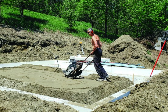 Prepping for the slab. After putting down horizontal wings of insulation around the base of the forms, the excavator backfills around the forms to lock them in place. Soil added inside the forms is compacted.