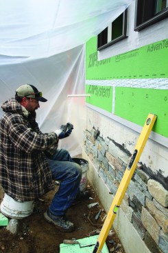 The blend of ledgestone and slightly larger ashlar stone comes with a nominal thickness of 1 1/2 inches. After trimming each stone to fit, the mason applies mortar to the bare concrete. He then butters the back of the stone ...