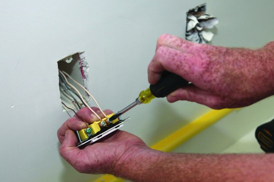 Holding the receptacle firmly in his hand, the author slips the loops for the neutral (white) conductors around the silver-colored screws and snugs the screws by hand.