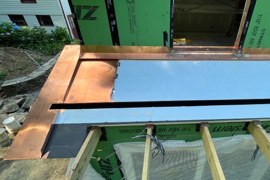 After installing high-temperature underlayment, the upslope flashing was installed. Copper windowsill pan flashing is soldered to continuous apron flashing at the roof-to-wall junction. Standing-seam panels are locked into the apron flashing and drip-edge flashing along the upslope edge of the built-in gutter.