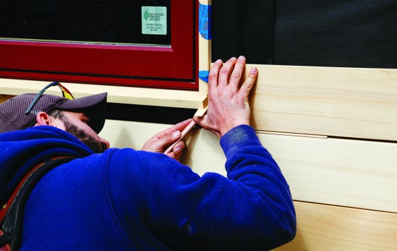 A carpenter scribes a board around a window in an open rainscreen system, with spacing between the boards and a space left under the window sill.