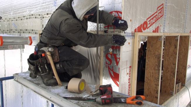 A carpenter applies 3M 8067 tape to the outer drainage-plane layer of rigid foam insulation. The crew accomplished the wall build-out during the severe winter of 2014 despite bitter cold and snow depths of 5 or 6 feet.