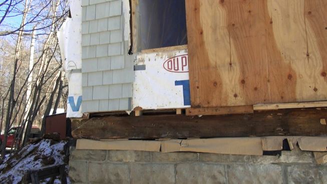 A view of the juncture between the existing foundation and wood frame, showing the existing siding, Tyvek housewrap, and sawn wood sill timber. The crew has already jacked up the sill a few inches in this view, in order to apply a Grace bituminous and polyethylene seal sealer.