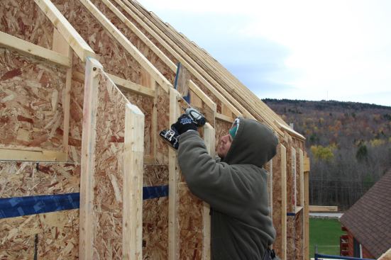 The carpenter sends a screw home from the wall I-joist flange into the roof I-joist flange. Roof sheathing will come down across this joint and terminate at the outboard face of the wall I-joists.