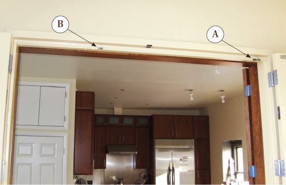 A closing coordinator mounted below the stop on the head jamb manages the two doors' closing sequence. The lever for the active, astragal-equipped door is mounted close to the side jamb (A); it holds the active door open until the passive door closes and activates a trigger mechanism located toward the center of the head jamb (B).