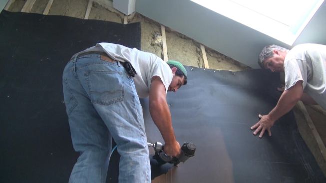 The crew fastens the Sound Barrier HD to the wall studs using a coil nailer loaded with one-inch roofing nails.