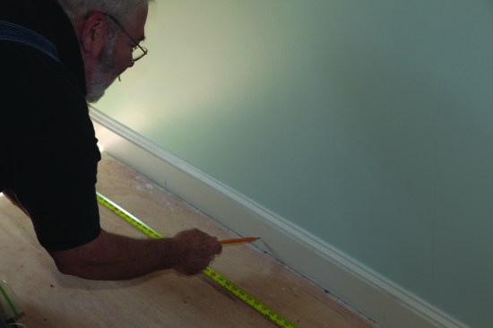 The recently painted baseboard stayed in place for this project, so the flooring butted against it. Before starting, the author lightly marked the joist locations on the baseboard for secure nailing. 
