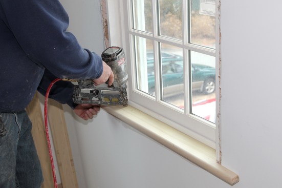The carpenter drives finish nails to fasten the stool to the sill frame of the window.