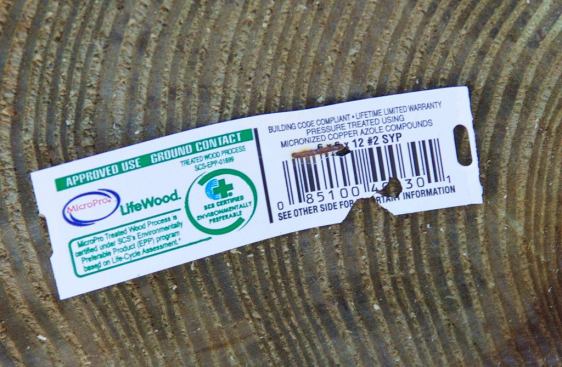 A close-up of the code-required label stapled to the end of a preservative-wood 6x6 Southern Yellow Pine post. The label shows the approved use (“ground contact”), and identifies the treatment chemical (“copper azole”).