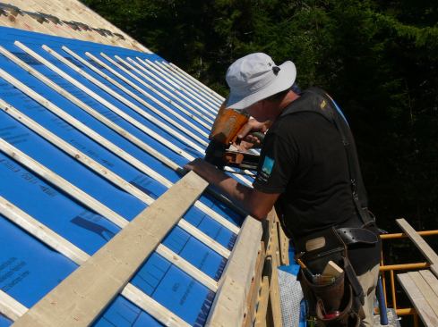 A carpenter applies horizontal 2x4 strapping to the roof over the 1x3 vertical strapping. This creates a space allowing air movement in all directions. Solitex Mento membrane is air-tight but highly vapor-open. The insulated space beneath the membrane will be able to dry into the vented air space created by the strapping. The horizontal 2x4 strapping will provide nailing for the metal roof, to be installed next.