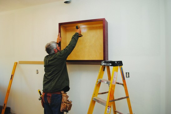 After marking the layout on the cleat, he positions the first cabinet and fastens it to the wall using screws.