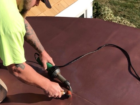 With membrane laid out and cut to the roof’s shape, the “field” seam work was welded first. Welding PVC is straightforward, though it takes some practice. First, the installer back-welds the seam by inserting the Leister torch nozzle about 2 inches under the lapped membrane (heating for a wider weld connection), while immediately pressing the pieces together using a seam roller, rolling 90 degrees to seam. On the outside edge, the seams are briefly heated by slipping a torch nozzle under the membrane edge at 45-degree angle, and then immediately pressed together using a seam roller, rolling at a 45-degree angle. The resulting weld is permanent and waterproof—it’ll never come apart again.