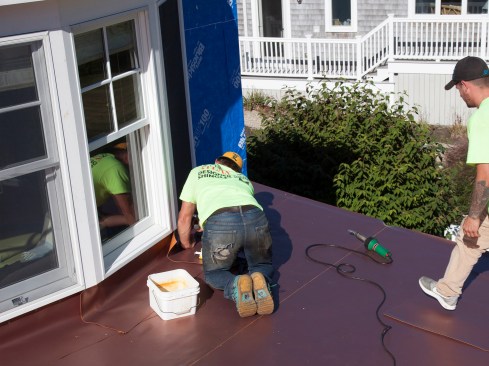 At roof-to-wall locations, flashing strips are cut from the roll, according to need. The strips are run “long” on to roof, then trimmed to length, later. When installing, the author’s crew places a few tack-welds on the flashing’s horizontal leg to hold the strip in place pending full seam welding.