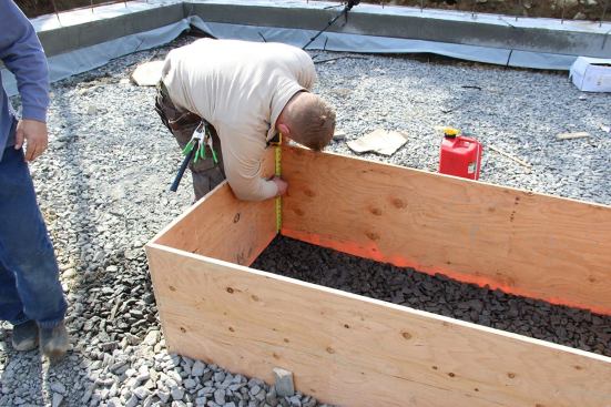 Carpenter Tim lays out the planned finish elevation for the concrete pad footing onto the plywood footing form, in preparation for placing base material and fitting insulation into the form.