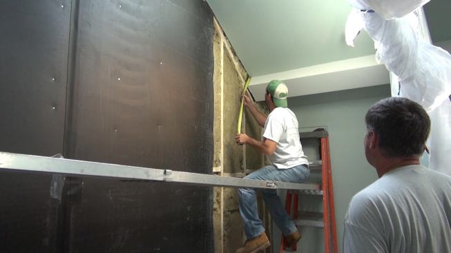 The crew measures a drop beam in the ceiling. The sound-deadening material has to be cut to fit any irregularities where the wall meets the ceiling.