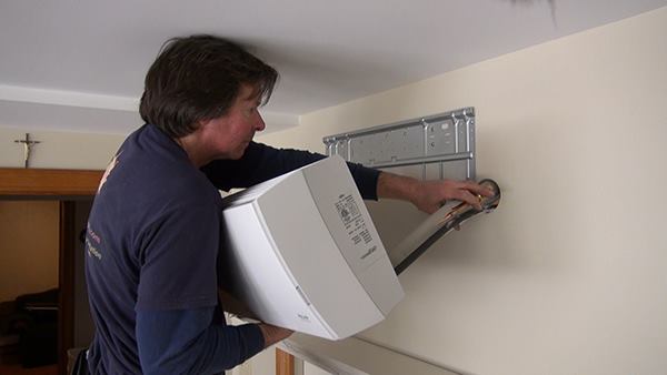 Chris Blaisdell threads the refrigerant lines, condensate tubing, and wiring cable for the system through a hole drilled in the partition wall where the indoor fan unit for the system will mount.