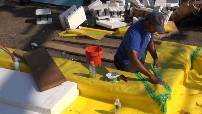 After gravel is placed and compacted, foam insulation and a poly vapor barrier are placed on the sub-base in preparation for placing the interior concrete slab floor. Here, Scotland Willis tapes a seam in the vapor barrier plastic.