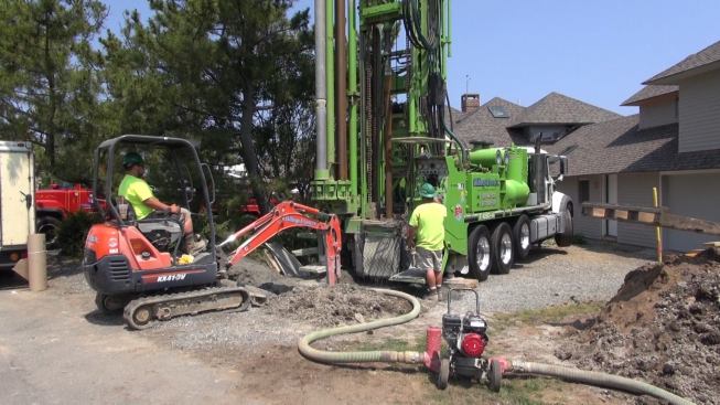 A well drilling rig from Amherst, New Hampshire-based 
Skillings and Sons drills one of three 400-foot-deep vertical wells for the geothermal ground loop for a ground source heat pump (GSHP) heating and cooling system for a house on the Massachusetts shore north of Boston. Drilling three wells took the crew and rig two days on site. The truck carries sections of drill shaft in a rack; as the well gets deeper, the operators pause to attach new drill sections to the shaft. In the foreground, a water pump extracts groundwater flowing from the borehole.