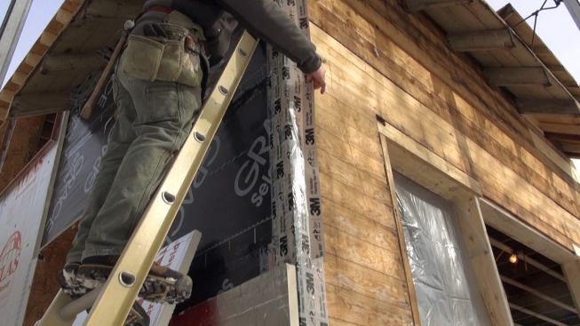 A worker applies 3m 8067 tape to an Ice and Water Shield edge during winter work on the exterior wall build-out.