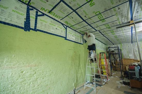 Here, the roof framing overhead has been covered with ProClima Intello smart vapor control membrane, and workers are taping the seams in the membrane using ProClima Tescon Vana tape. The membrane is also being taped to the Sto Emerald Cote surface of the brick party wall. The long brick wall divides the residence from the conditioned space of an adjoining neighbor, and therefore does not have to be insulated.